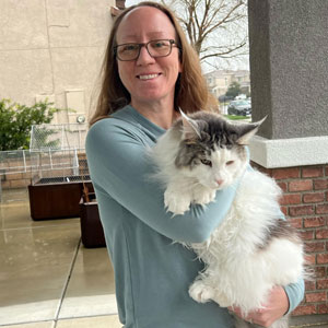 A smiling veterinarian with glasses and long hair holds a large, fluffy white and gray cat outdoors near a brick wall on a rainy day.