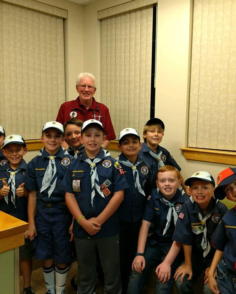 A group of young scouts in uniform pose and smile for a photo indoors with an older man, who is a local vet, standing behind them. All wear blue shirts, neckerchiefs, and caps. They appear happy and are gathered closely together.