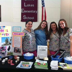 Four women stand and smile behind a table with educational materials, dental care items, and cat-themed resources at a school event. A “Great Lakes Elementary School” banner and an American flag are visible in the background.
