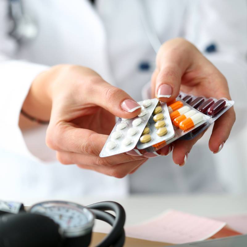 A veterinarian in a white coat holds several blister packs of various colored pills, with medical equipment visible in the foreground.